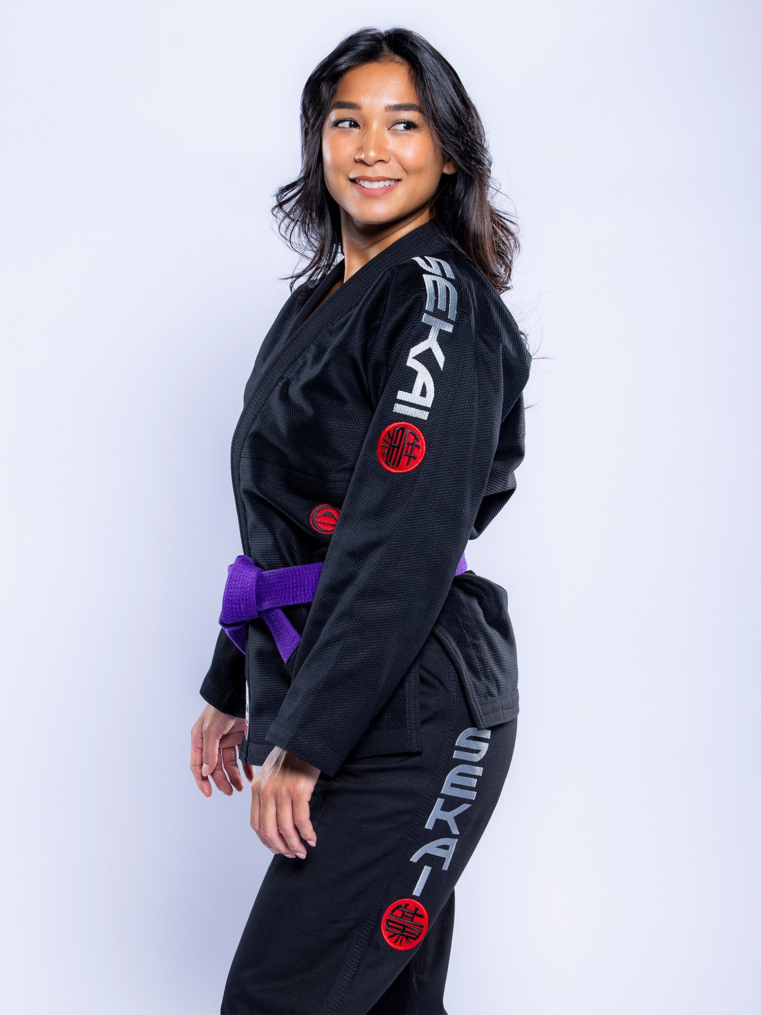 A woman in a black and grey Womens SEKAI 3.0 BJJ Gi with red patches and a purple belt stands sideways, smiling against a plain light background.