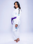 A woman stands barefoot on a white background, smiling and facing sideways with her head turned toward the camera. She is wearing the Womens SEKAI 3.0 BJJ Gi White Teal, featuring a purple belt and black and blue patches.