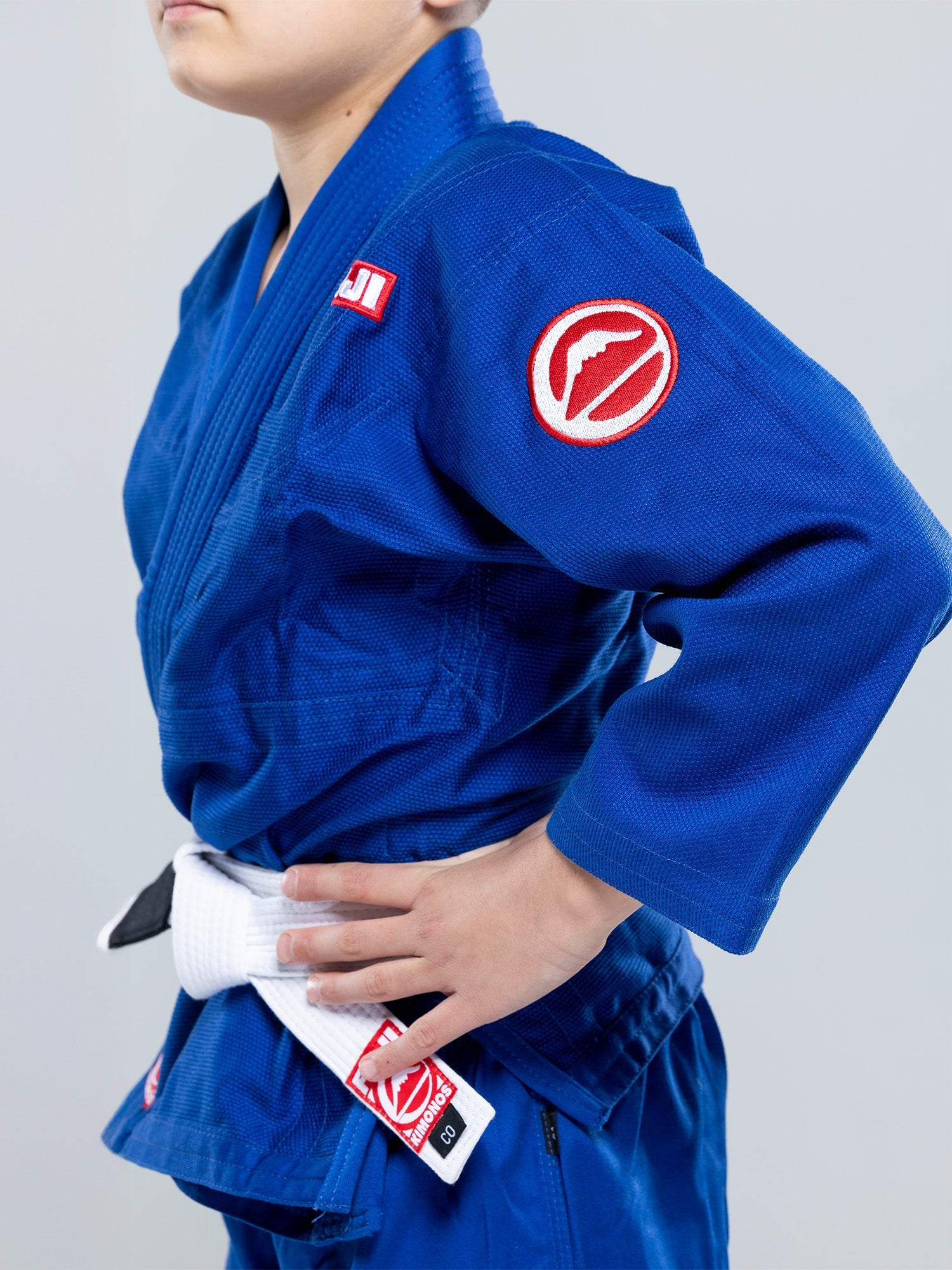 A child wearing the Kids Competition BJJ Gi Blue with a white belt stands hand on hip. The gi features red and white patches on the arm and waist. The background is plain and light-colored.
