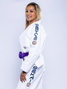 A woman in the Womens SEKAI 3.0 BJJ Gi White Navy, featuring sleeve branding and patches, wears a purple belt and smiles in side profile against a plain white backdrop.