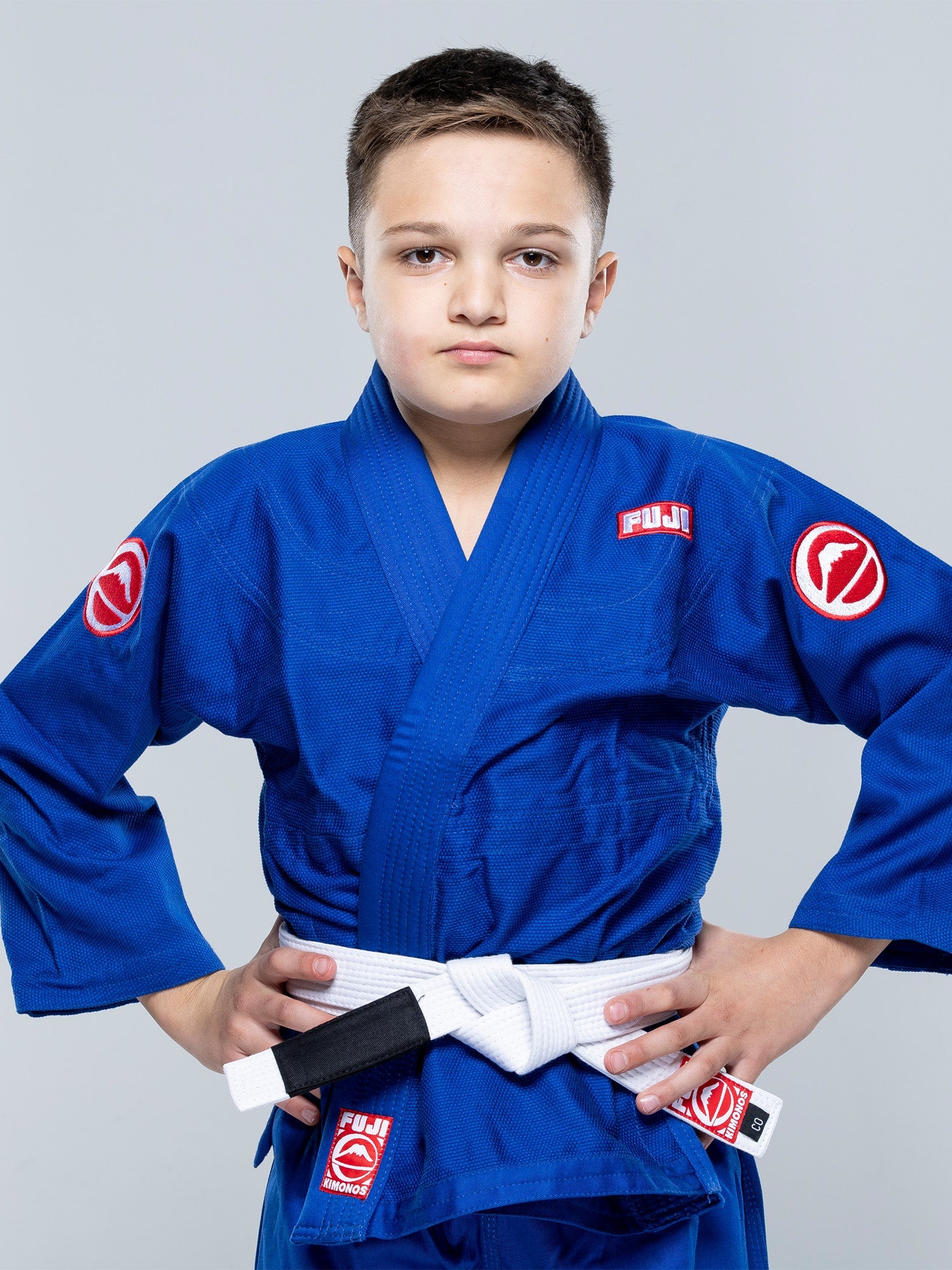 A young boy wears the Kids Competition BJJ Gi Blue with a white belt, standing confidently with his hands on his hips against a plain light gray background.