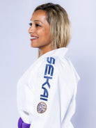 A blonde woman in profile smiles while wearing the Womens SEKAI 3.0 BJJ Gi White Navy with a purple belt, "SEKAI" visible on the sleeve, against a plain light background.