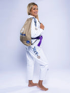 A woman smiles barefoot in the Womens SEKAI 3.0 BJJ Gi White Navy with a purple belt, carrying a tan drawstring bag on her back and posing against a plain light background.
