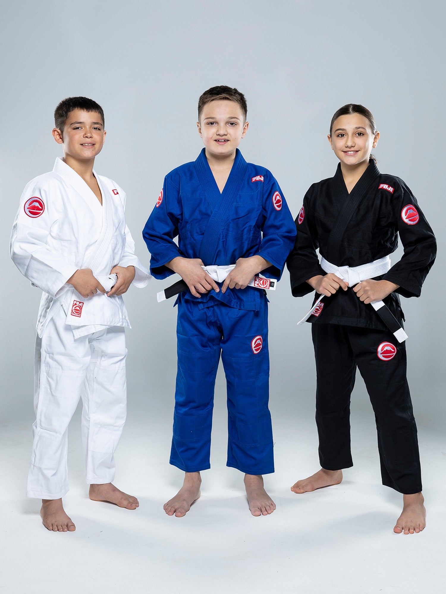 Three children in martial arts uniforms stand smiling. The child in the center wears the Kids Competition BJJ Gi Blue, while the others wear white and black uniforms. All are wearing white belts.