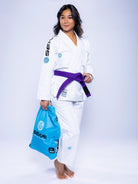 A woman wearing the Womens SEKAI 3.0 BJJ Gi White Teal with a purple belt stands barefoot on a light background, confidently smiling at the camera while holding a light blue bag.