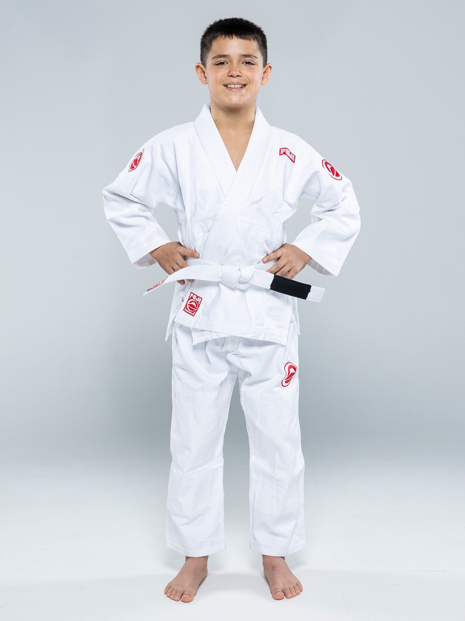 A smiling boy stands barefoot on a white floor, wearing the Kids Competition BJJ Gi White with a white belt, hands on his hips, against a plain light gray background.
