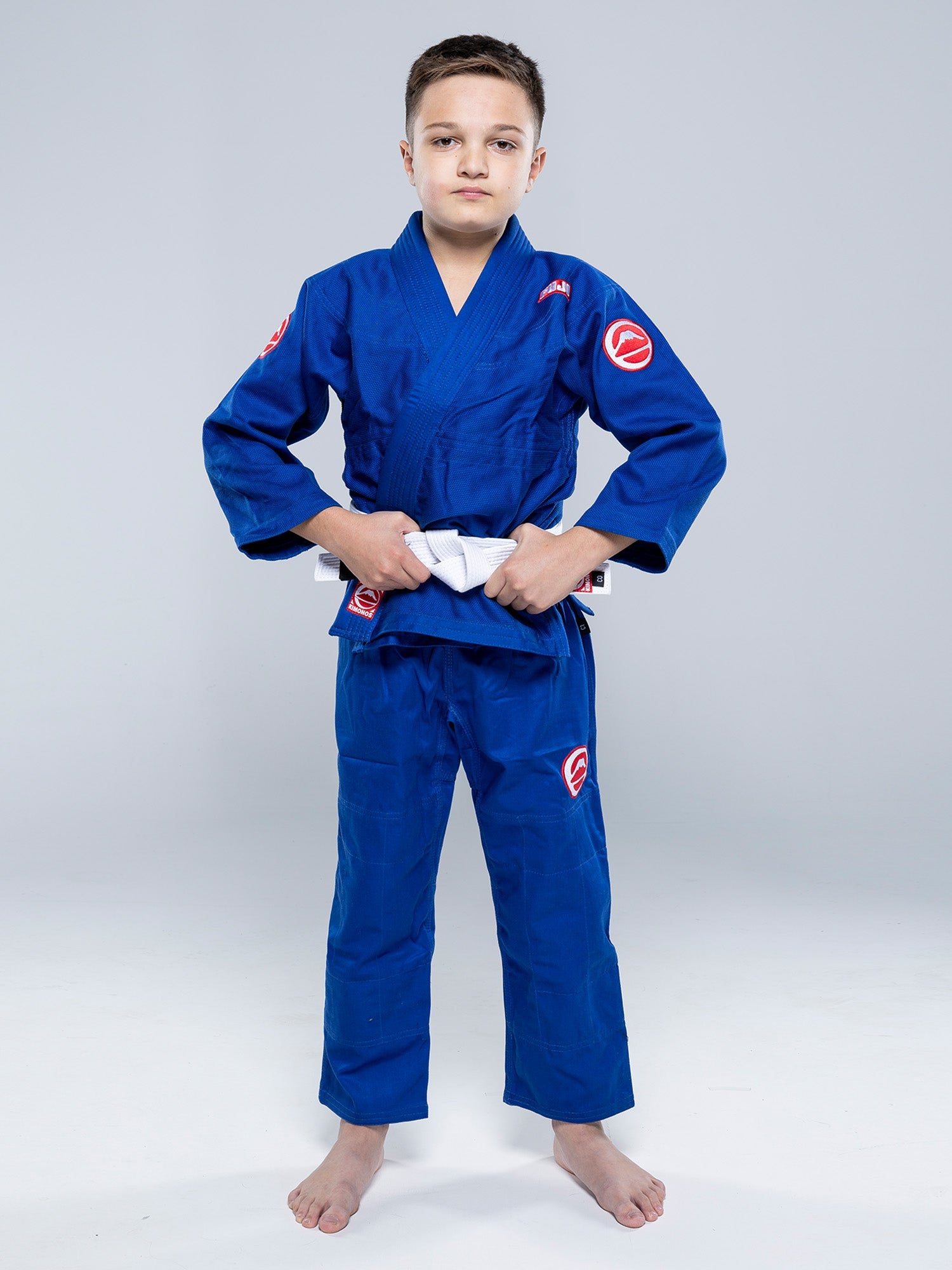 A young boy wears the Kids Competition BJJ Gi Blue, barefoot on a white floor, holding his white belt and facing the camera with a neutral expression.
