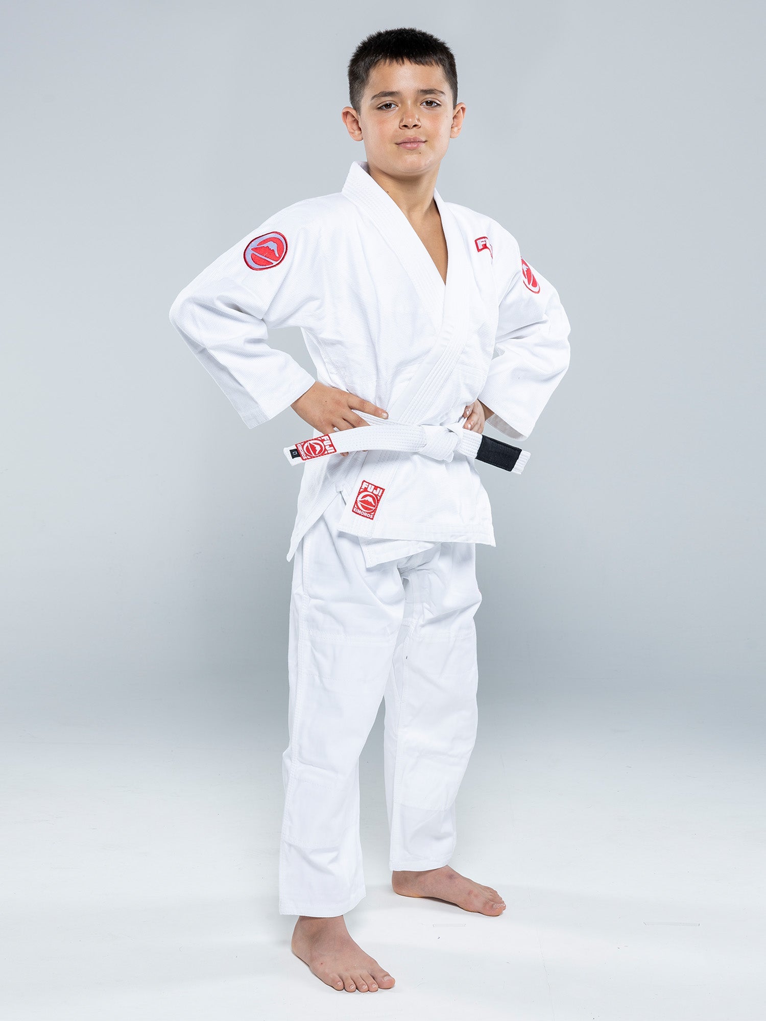 A young boy stands confidently in the Kids Competition BJJ Gi White with a white belt, barefoot, hands on hips, looking at the camera against a plain light gray background.