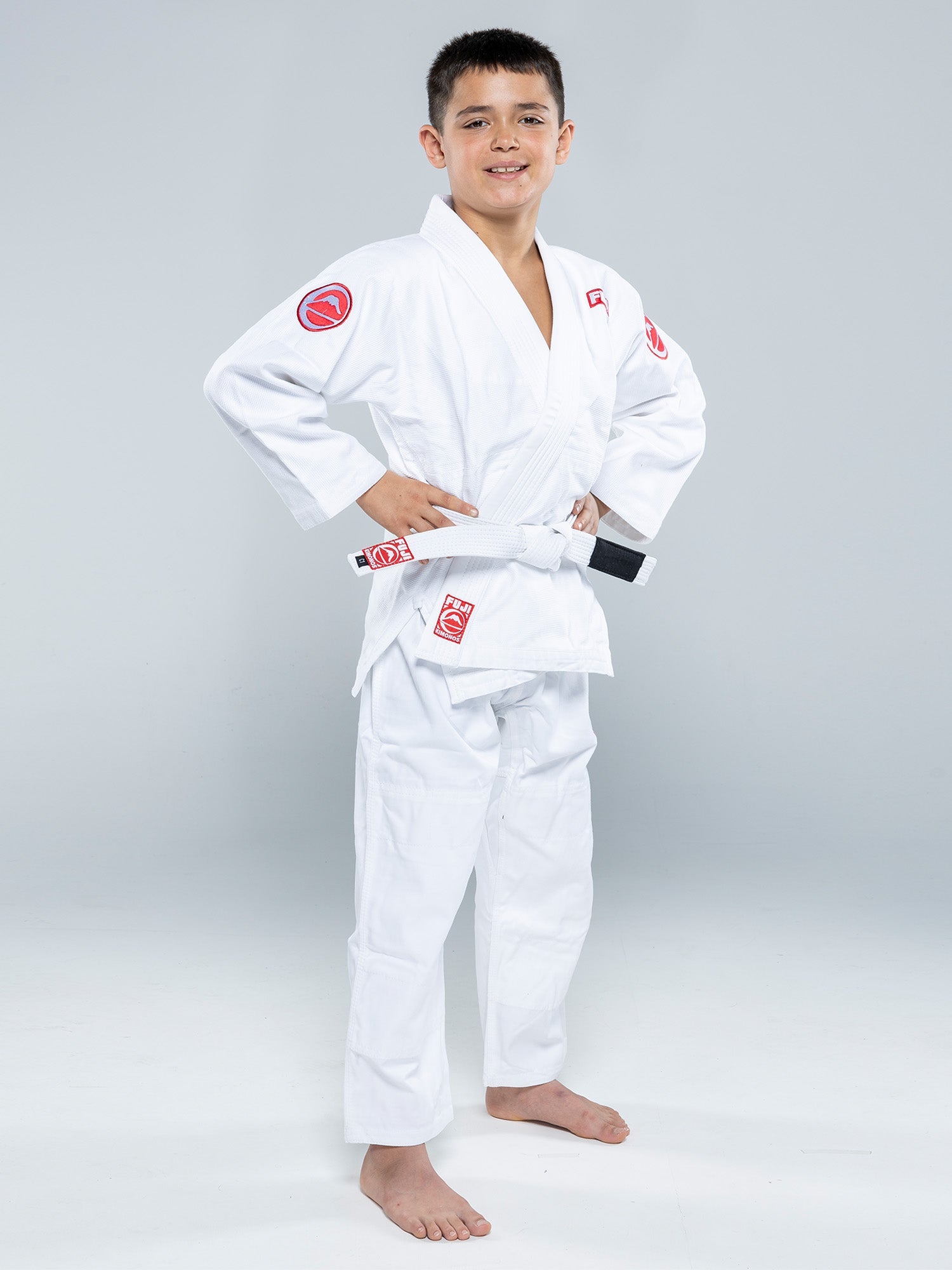 A young boy stands barefoot in a Kids Competition BJJ Gi White with a white belt, hands on hips and smiling at the camera against a plain gray background.
