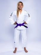 A woman stands barefoot on a light background, smiling with hands on hips. She is wearing the Womens SEKAI 3.0 BJJ Gi White Navy and a purple belt.