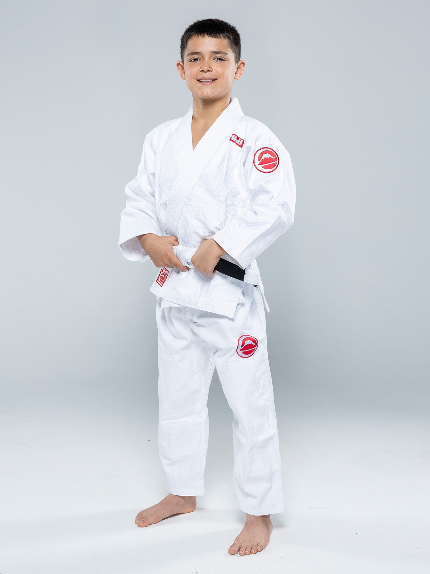 A boy stands barefoot on a white floor, smiling and holding his black belt while wearing the Kids Competition BJJ Gi White with red patches.