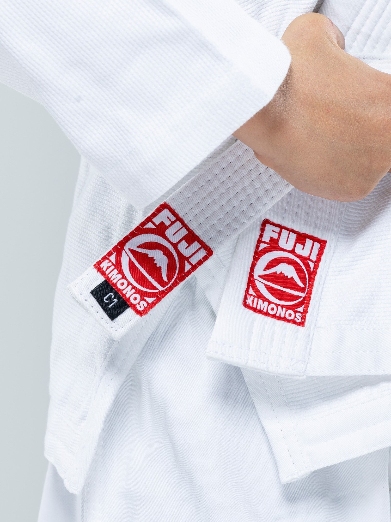 Close-up of a person wearing the Kids Competition BJJ Gi White with red FUJI KIMONOS patches and a black C1 tag on the sleeve and jacket, gripping the lapel.