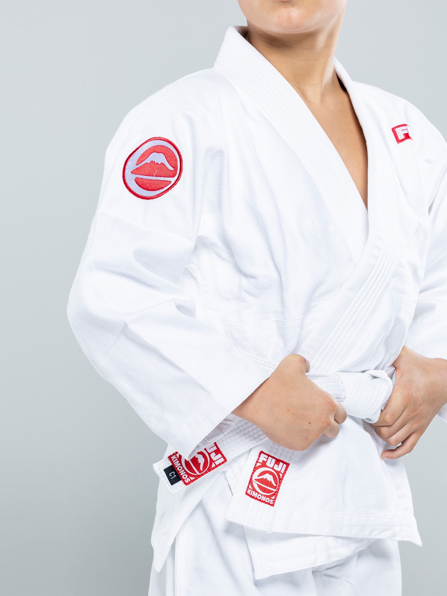 A child adjusts the belt on a Kids Competition BJJ Gi White, focusing on the upper body with hands tying the belt and embroidered logos visible on the white uniform.