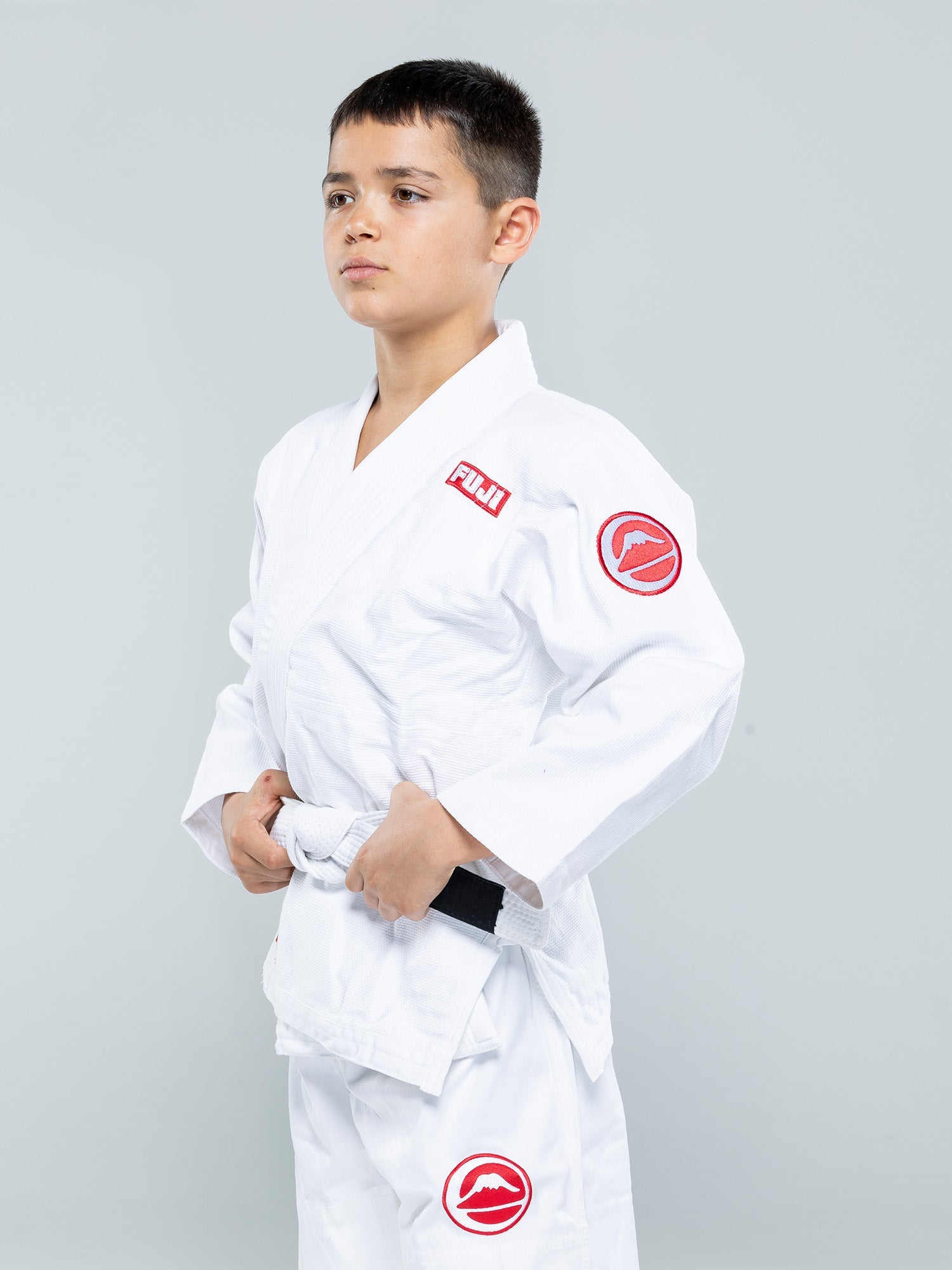 A young boy confidently models the Kids Competition BJJ Gi White, holding his white belt ends with both hands against a plain light gray background.