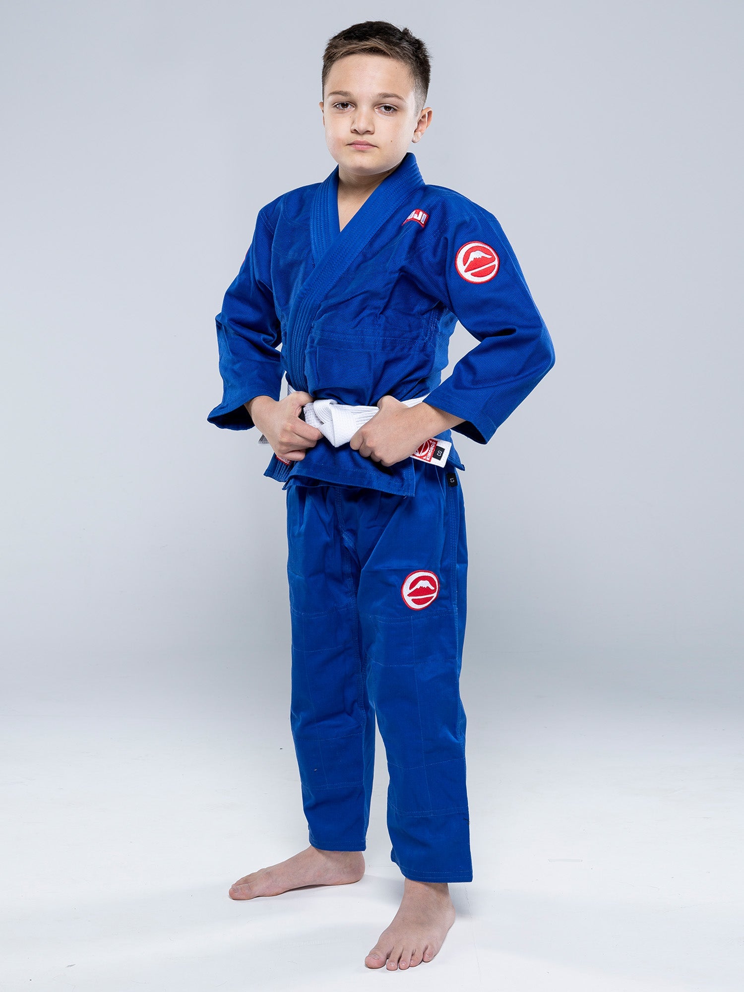 A young boy stands barefoot on a white floor, confidently looking at the camera with his hands on his belt, wearing the Kids Competition BJJ Gi Blue and a white belt.