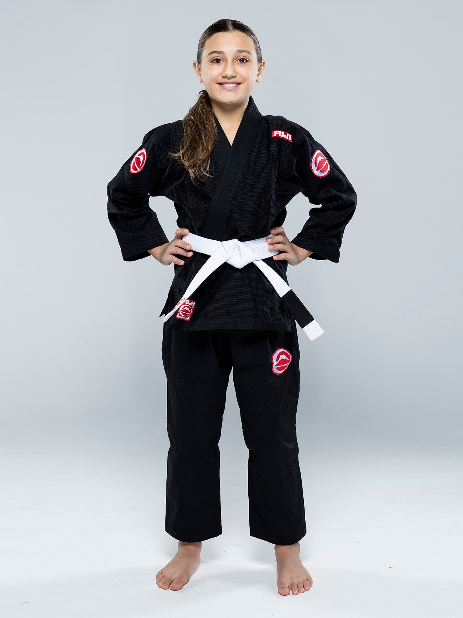 A young girl stands barefoot, smiling with hands on hips, wearing the Kids Competition BJJ Gi Black with a white belt and red patches, set against a plain gray background.