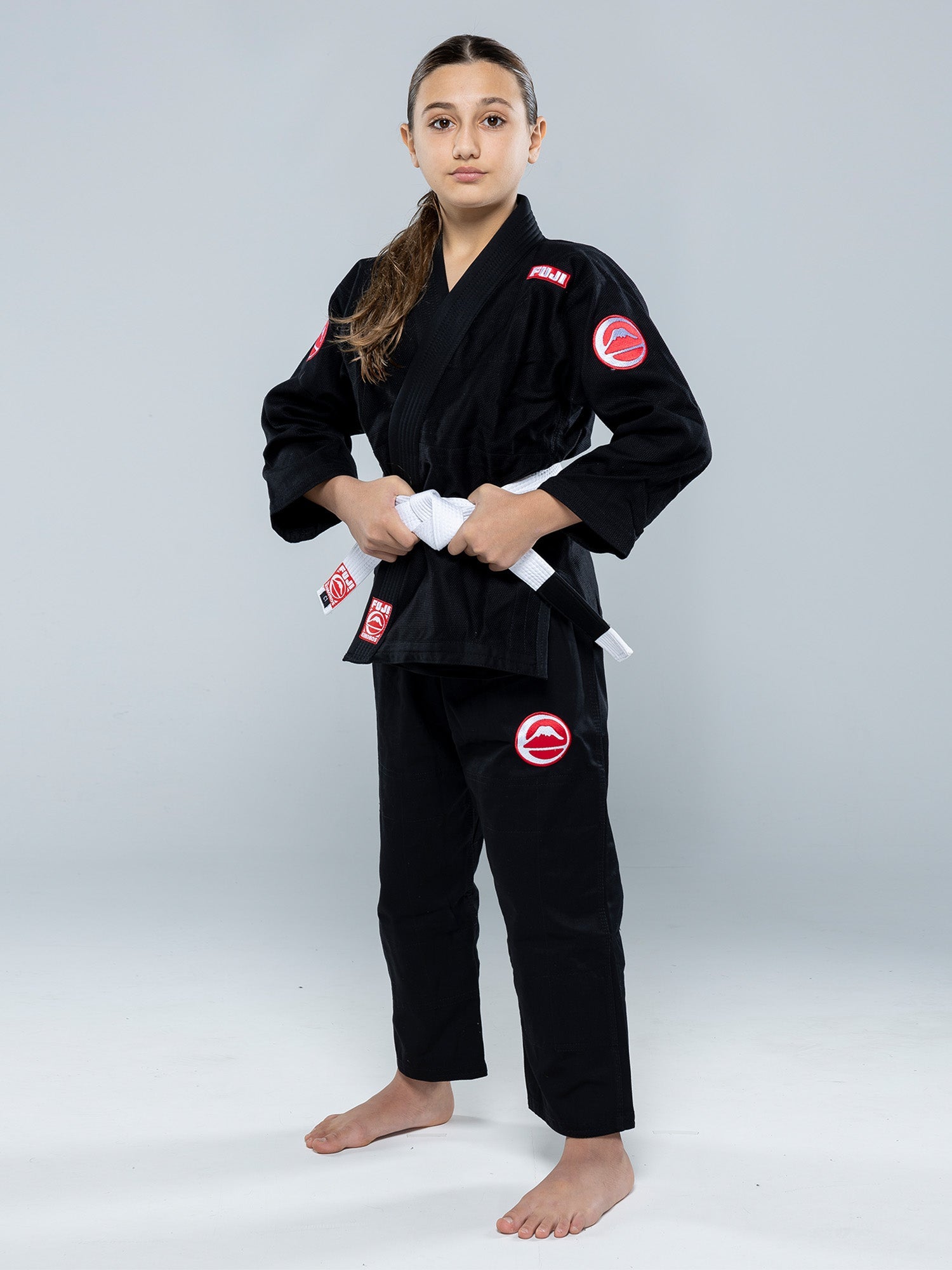 A young girl stands barefoot on a white floor, wearing the Kids Competition BJJ Gi Black and holding a white belt, looking confidently at the camera.