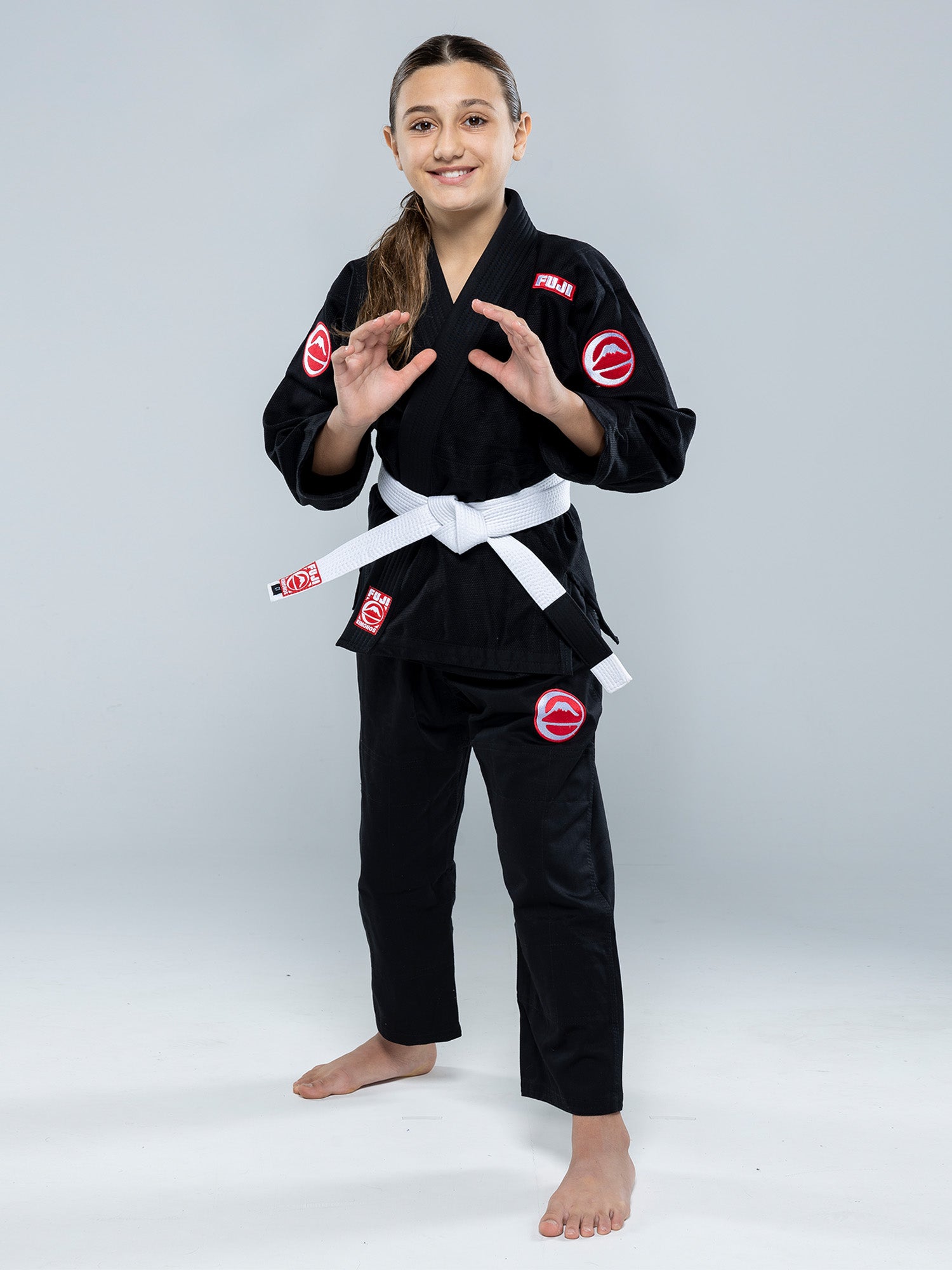 A young girl stands barefoot on a white floor, smiling and posing in a ready stance while wearing the Kids Competition BJJ Gi Black with a white belt and red and white patches.