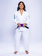 A woman with long dark hair, smiling and standing barefoot in the Womens SEKAI 3.0 BJJ Gi White Teal, holds a purple belt against a plain light background.