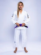 A woman stands barefoot against a light background, wearing the Womens SEKAI 3.0 BJJ Gi White Navy and holding a purple belt, facing forward with a neutral expression.