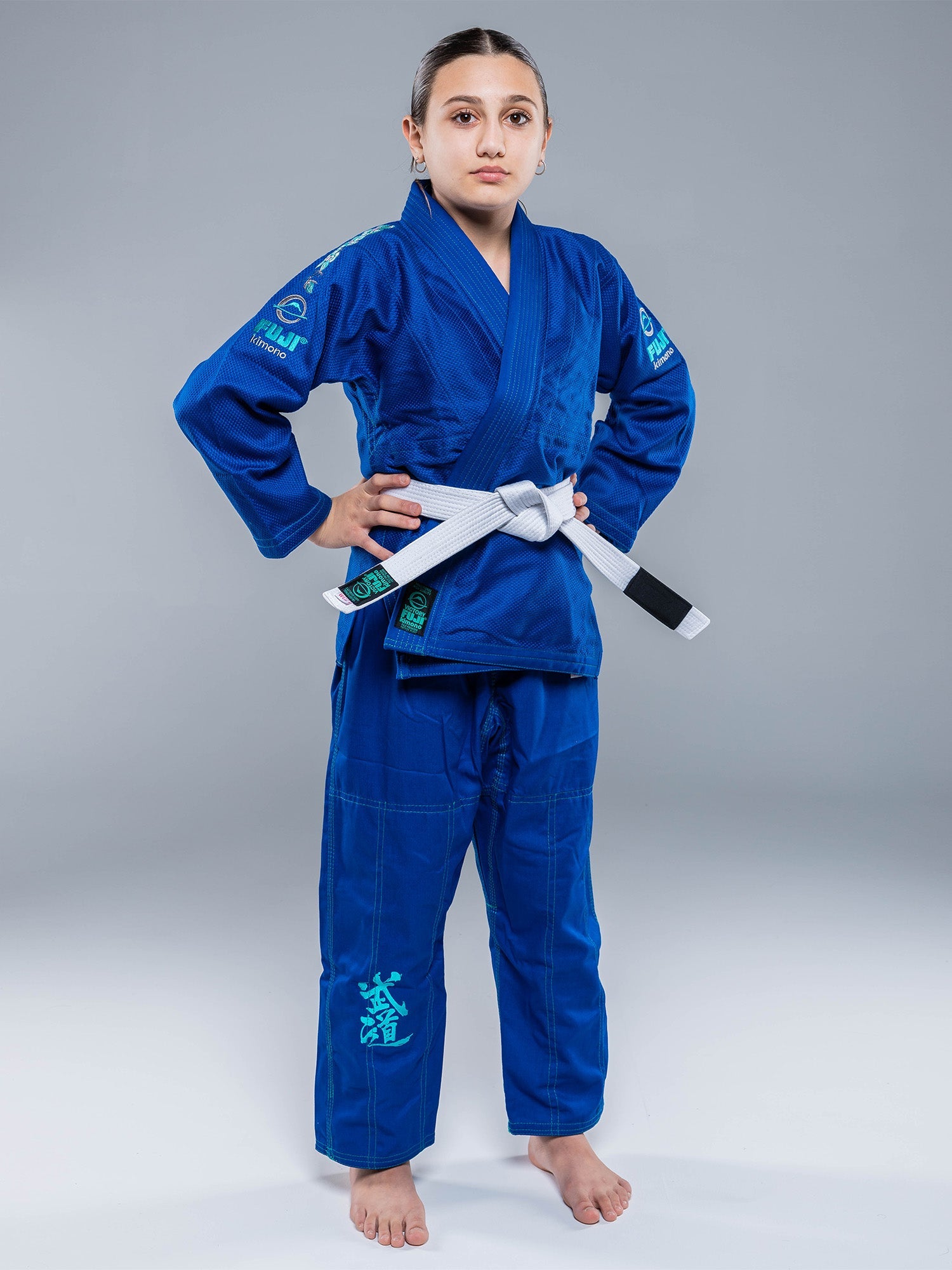 A young person stands confidently in the Blossom Kids Jiu Jitsu Gi Blue with a white belt, hands on hips, against a plain gray background.