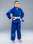 A young person stands confidently in the Blossom Kids Jiu Jitsu Gi Blue with a white belt, hands on hips, against a plain gray background.