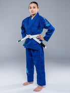 A young person wears the Blossom Kids Jiu Jitsu Gi Blue with a white belt, standing barefoot on a plain gray background, hands on hips and looking forward confidently.