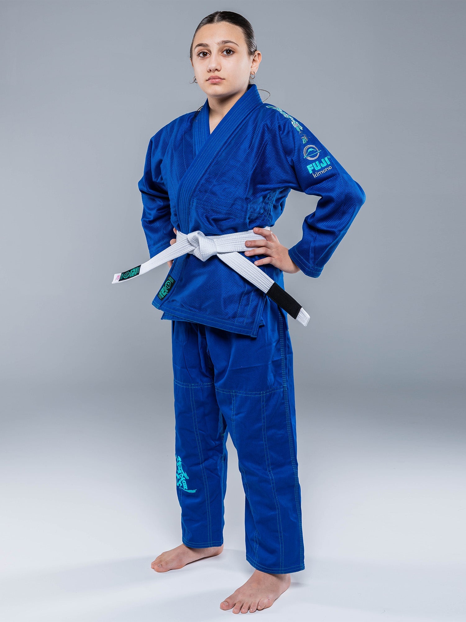 A young person wears the Blossom Kids Jiu Jitsu Gi Blue with a white belt, standing barefoot on a plain gray background, hands on hips and looking forward confidently.