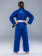 A youth wearing the Blossom Kids Jiu Jitsu Gi Blue with a white belt stands facing away, hands on hips, against a plain gray background.