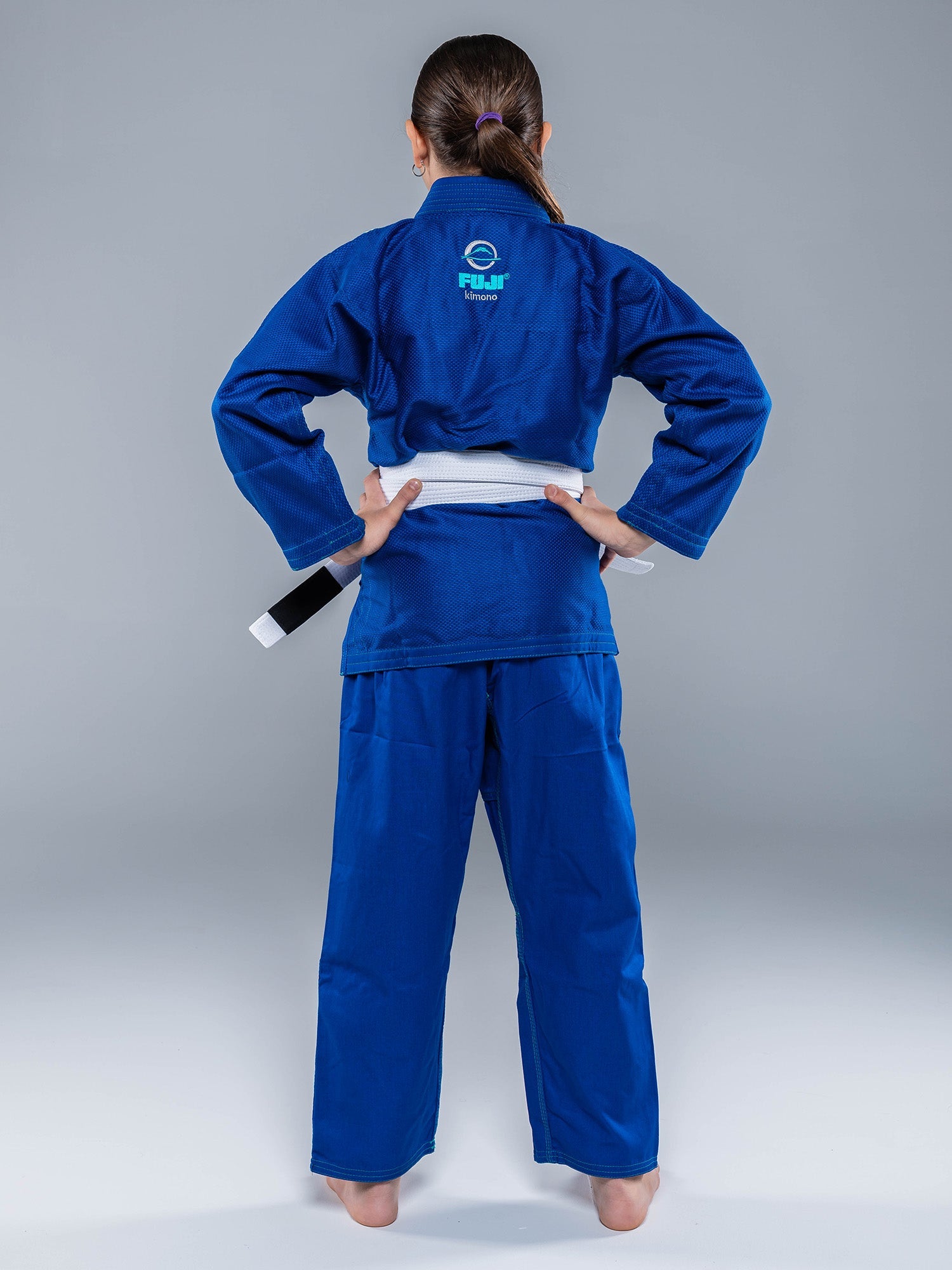 A youth wearing the Blossom Kids Jiu Jitsu Gi Blue with a white belt stands facing away, hands on hips, against a plain gray background.
