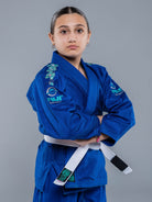 A young person stands confidently with arms crossed, wearing the Blossom Kids Jiu Jitsu Gi Blue featuring floral embroidery and a white belt, set against a plain gray background.