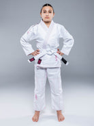 A young person, barefoot, wears the Blossom Kids Jiu Jitsu Gi White with a white belt and stands facing forward with a serious look against a plain gray background.