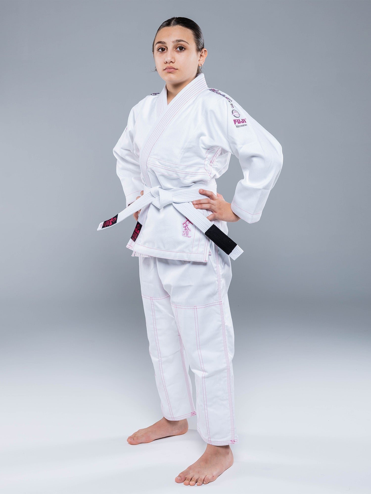 A person stands confidently in the Blossom Kids Jiu Jitsu Gi White with pink trim and a white belt, hands on hips and barefoot against a plain gray background.