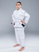 A person stands confidently in the Blossom Kids Jiu Jitsu Gi White with pink trim and a white belt, hands on hips and barefoot against a plain gray background.